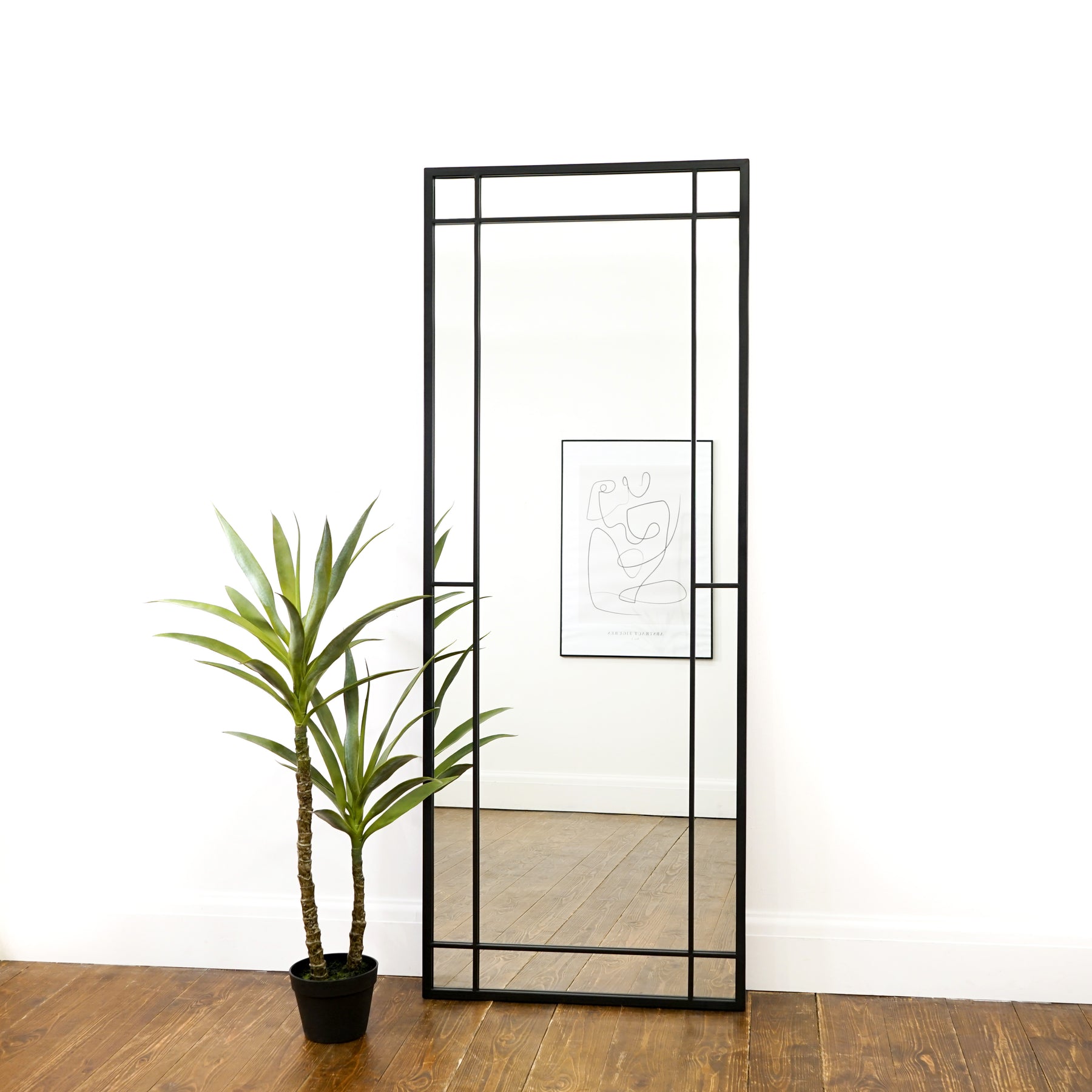 Black industrial full length metal mirror leaning against wall with plant